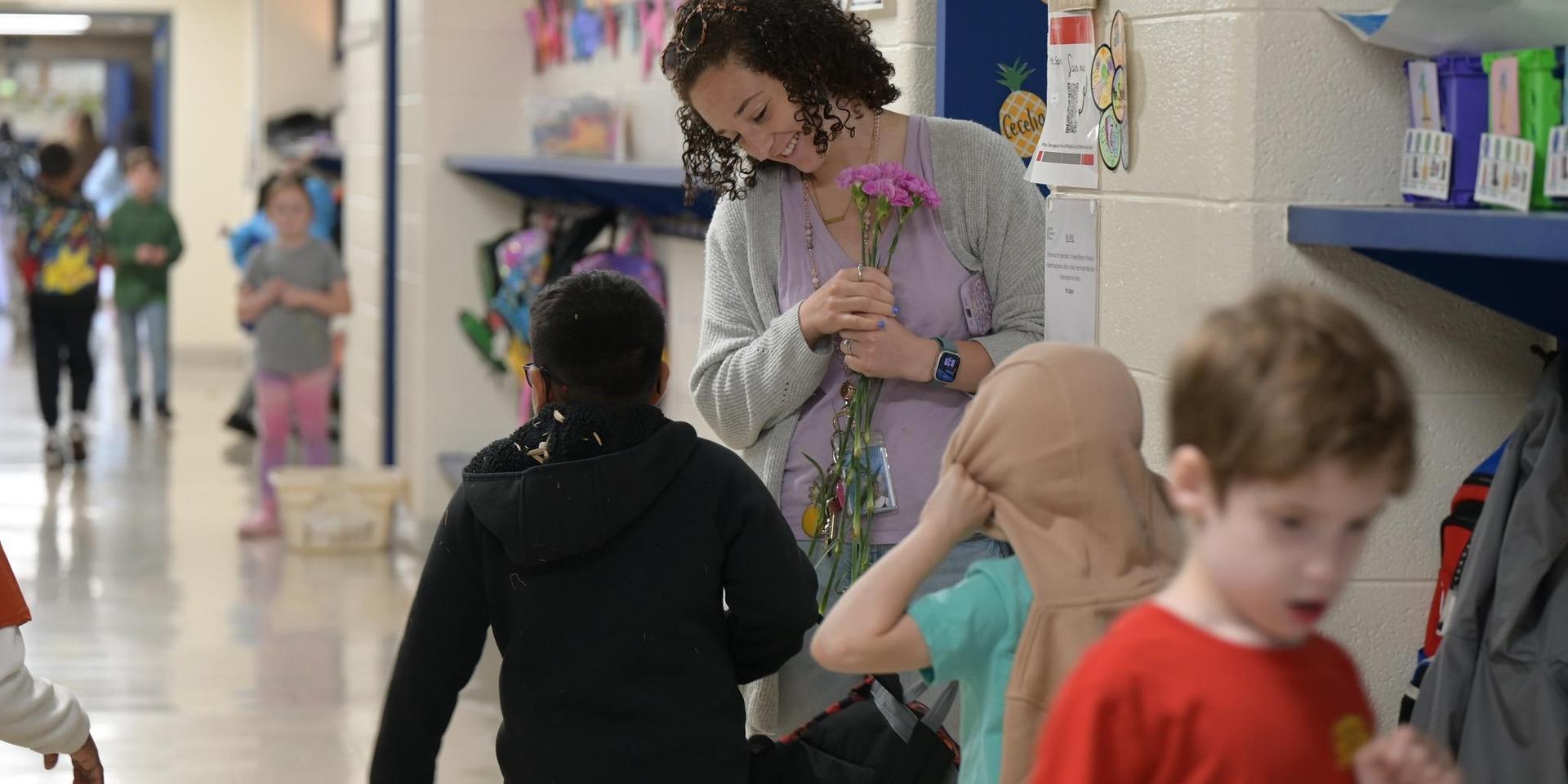 Teacher holding flowers in a busy school hallway with children walking by.