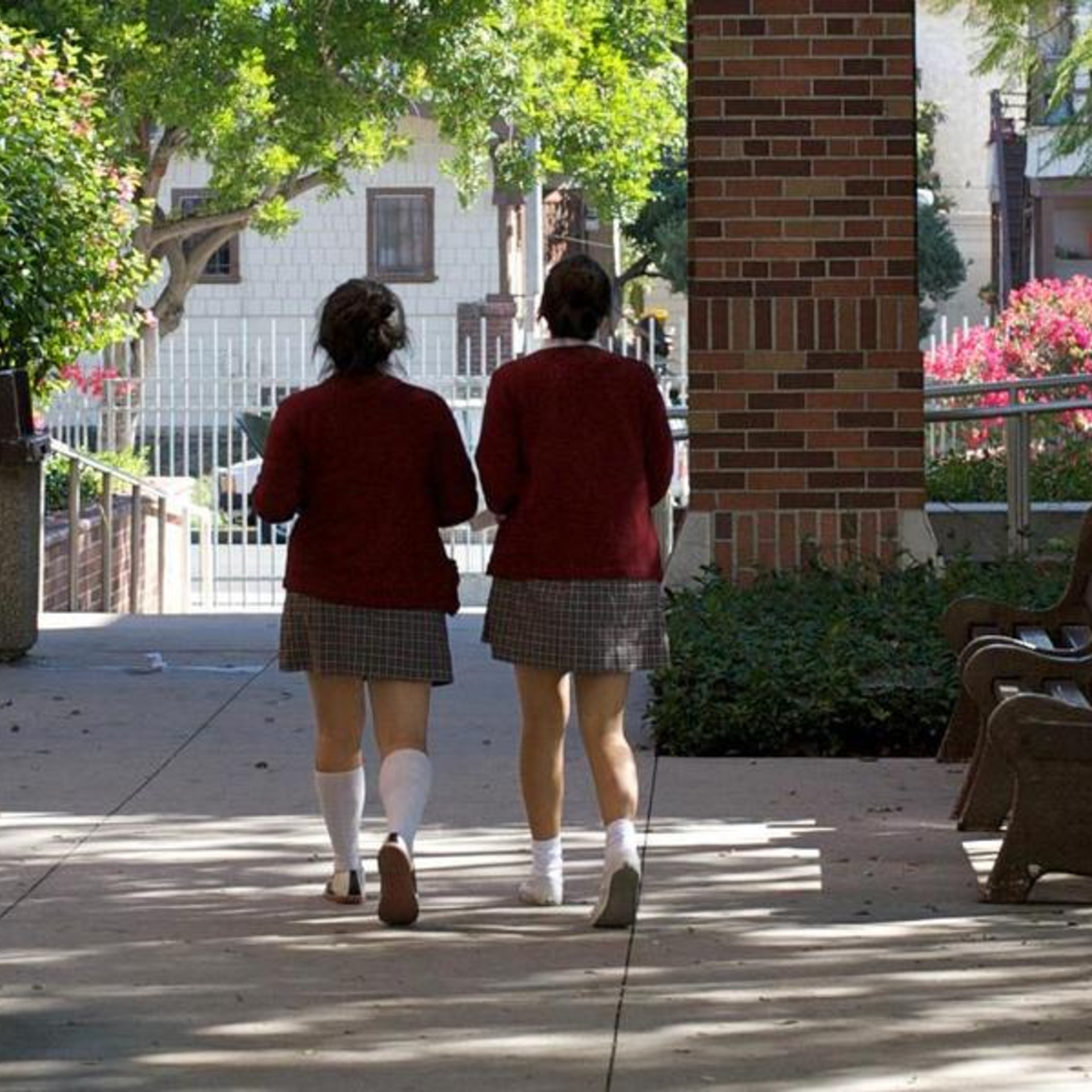 Two students in red sweaters and plaid skirts walking together down a pathway.