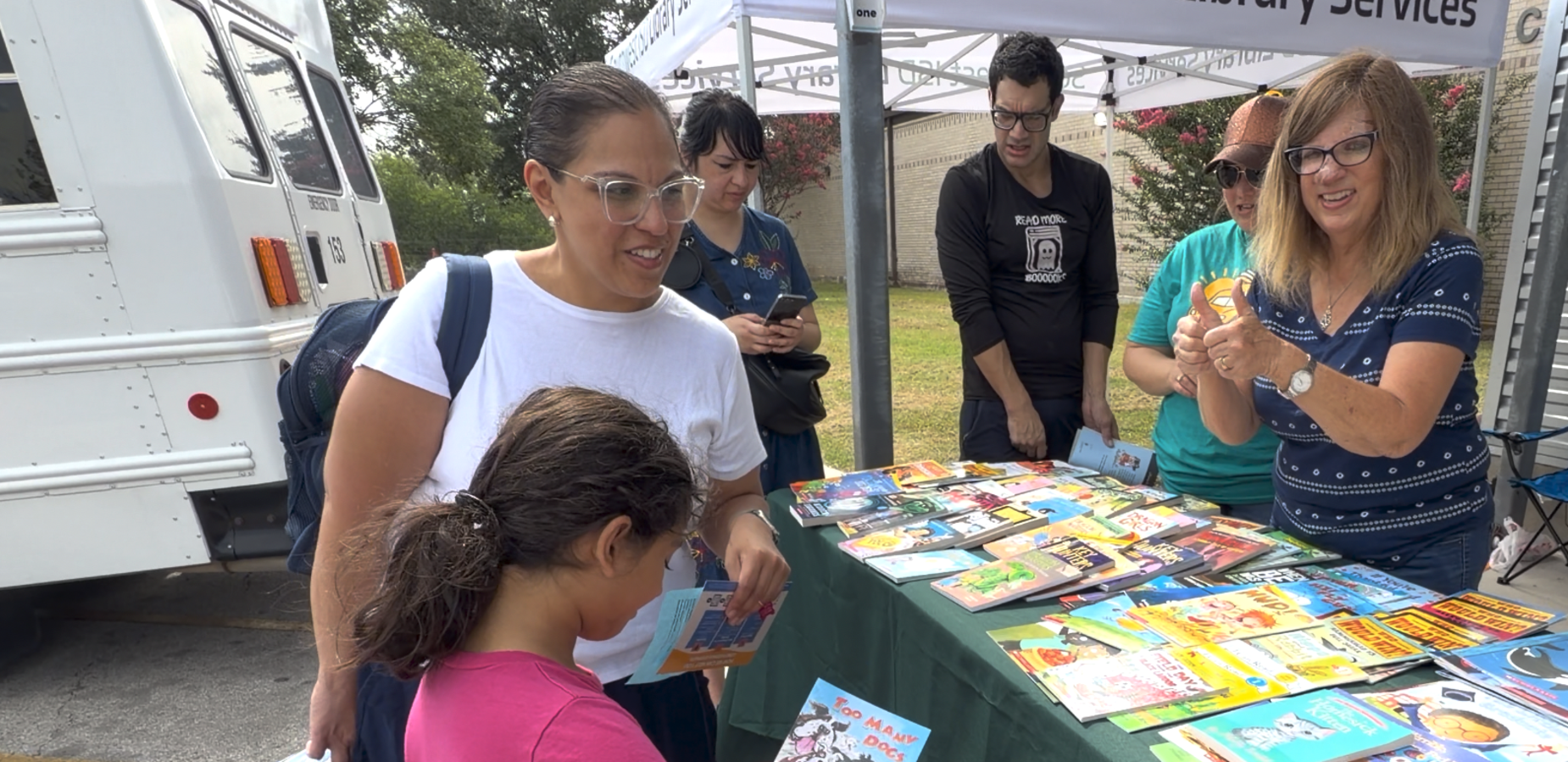 A group of people at an outdoor event, with children exploring books and a pumpkin.