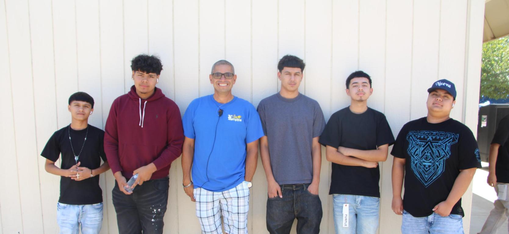 Six young men standing in a row against a light-colored wall.