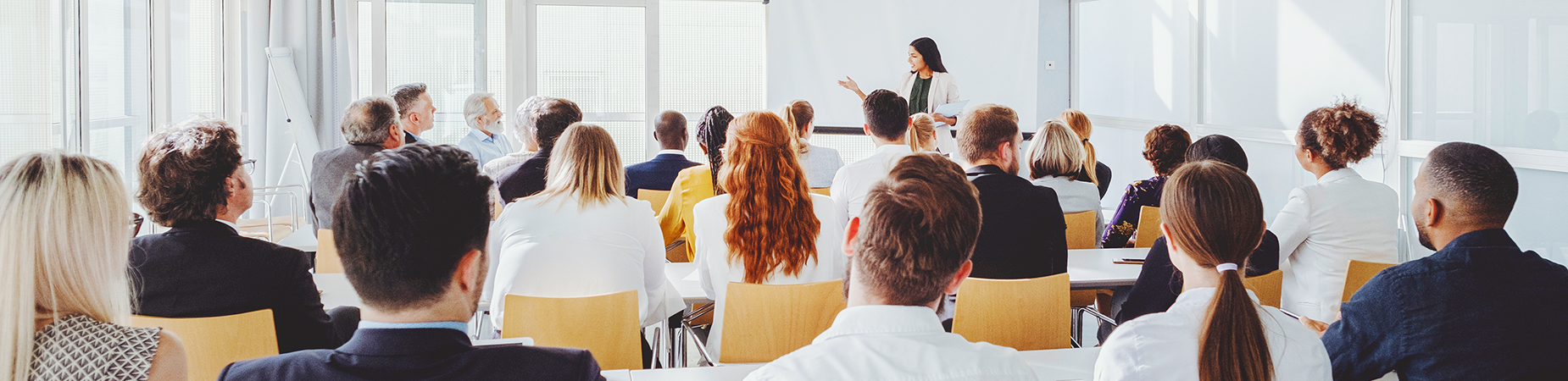 A training room filled with four or more rows of people watching a trainer. The people have their backs to the camera.
