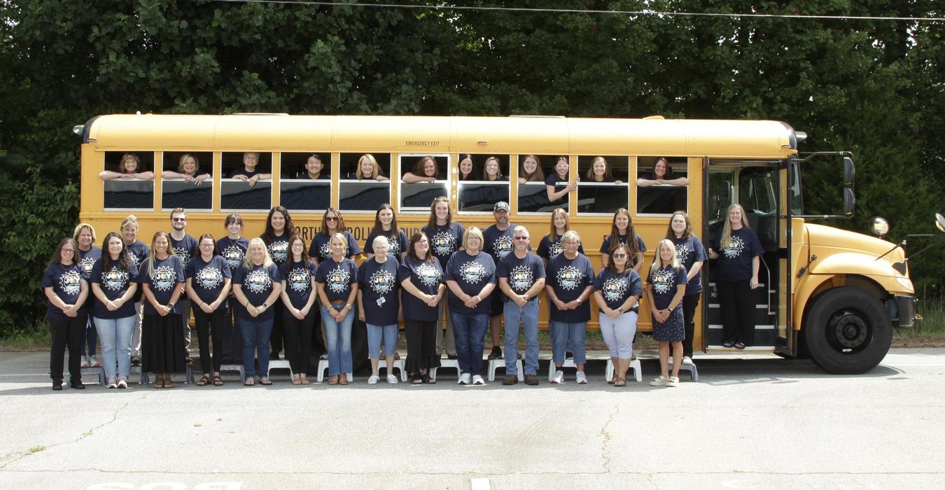 Group photo of staff members in matching shirts beside a yellow school bus.