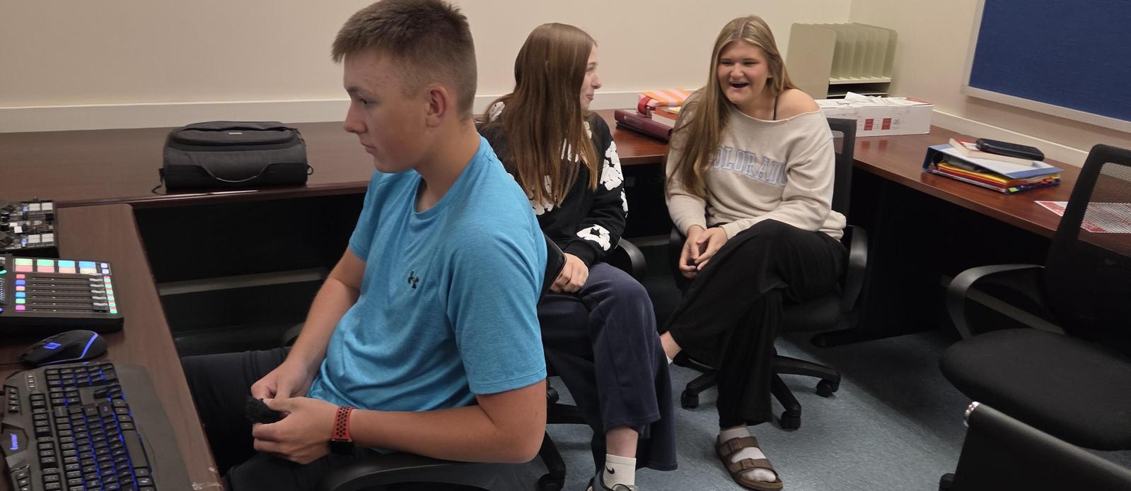Podcast production photo, a student sitting by the control panel and two students sitting in the background.
