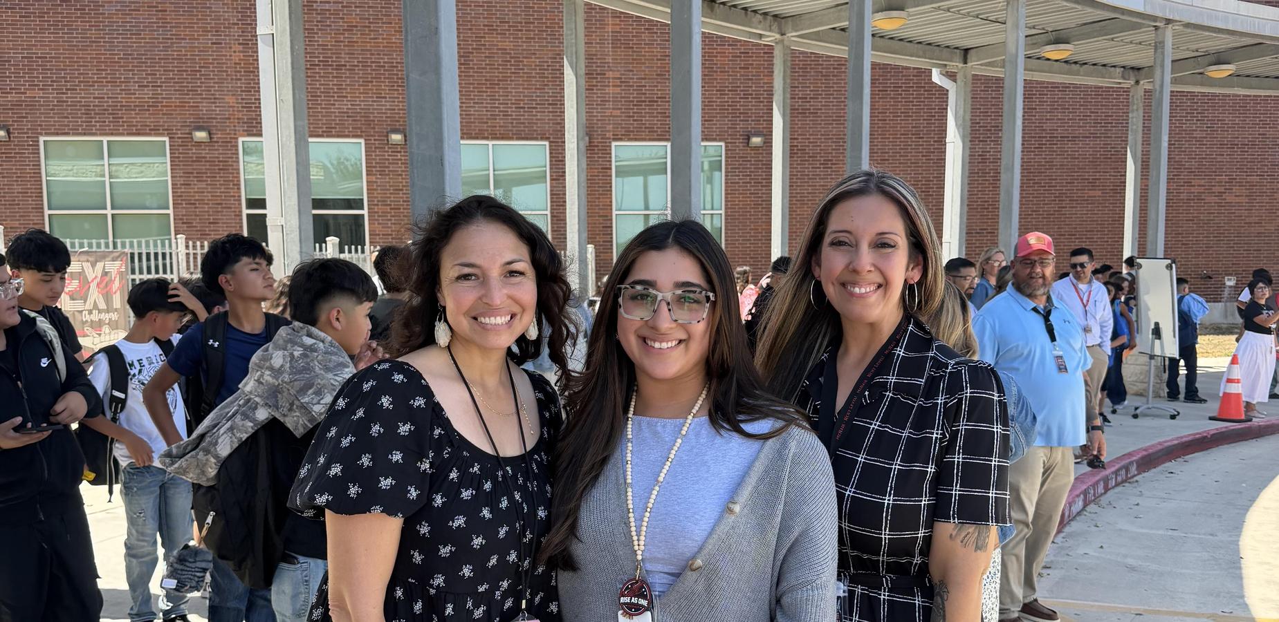 Three women pose together outdoors, smiling with a crowd in the background.