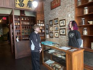 Two individuals stand in a historical setting with shelves displaying jars and framed images.