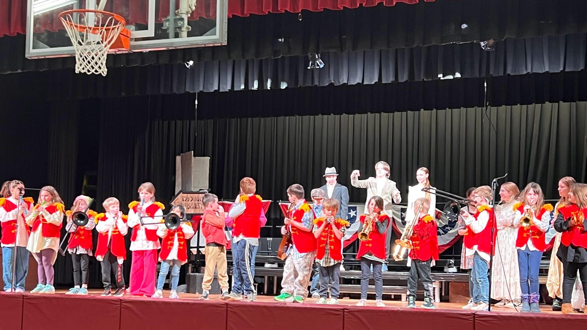 Children in red jackets performing with musical instruments on stage.