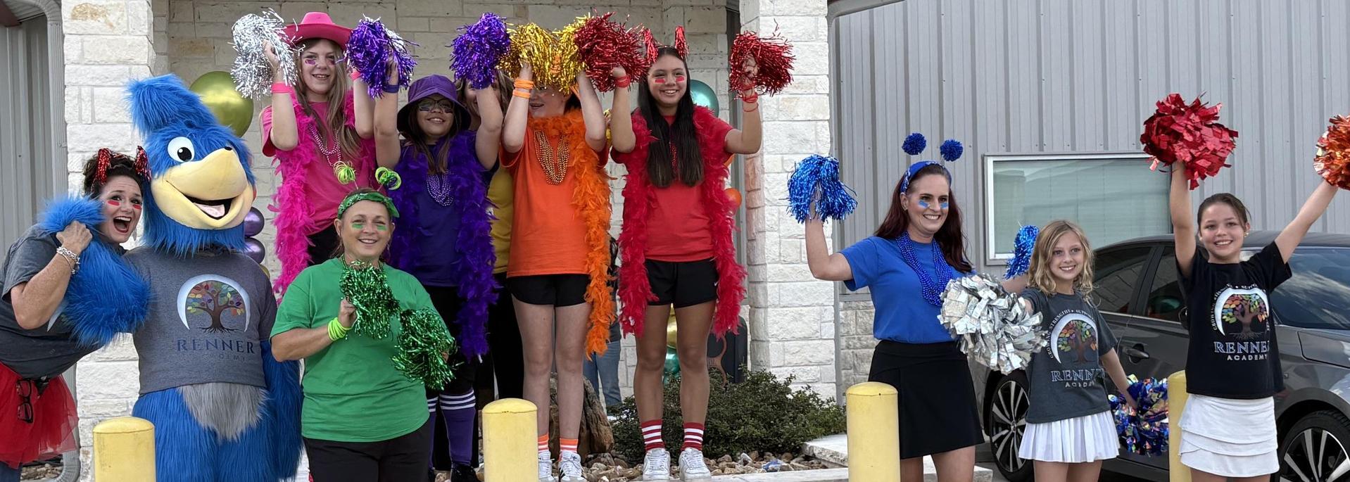 A group of cheerful people in bright outfits and a mascot pose with pom-poms outside.