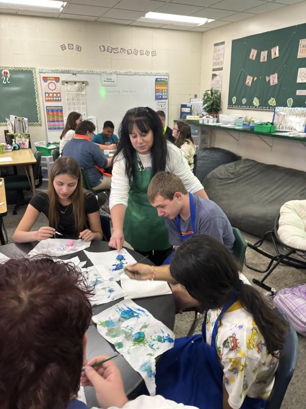 A teacher assisting students with painting at a classroom table.