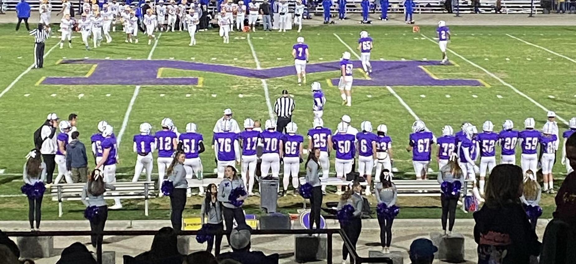A football team in purple uniforms watches the game on a grassy field with spectators.