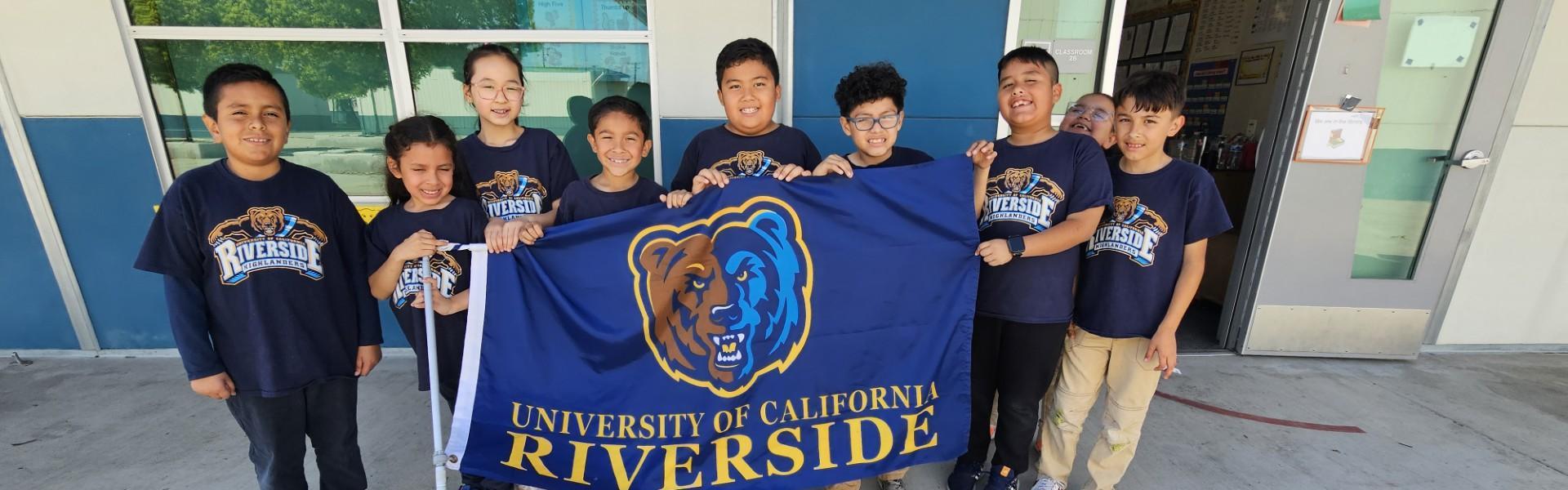 Group of children holding a University of California Riverside flag outdoors.