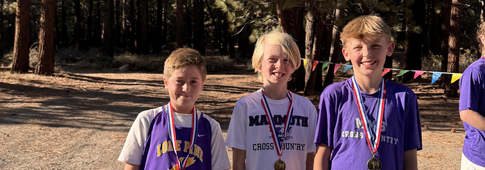 Three boys smiling with medals in a forested area, celebrating a cross country event.