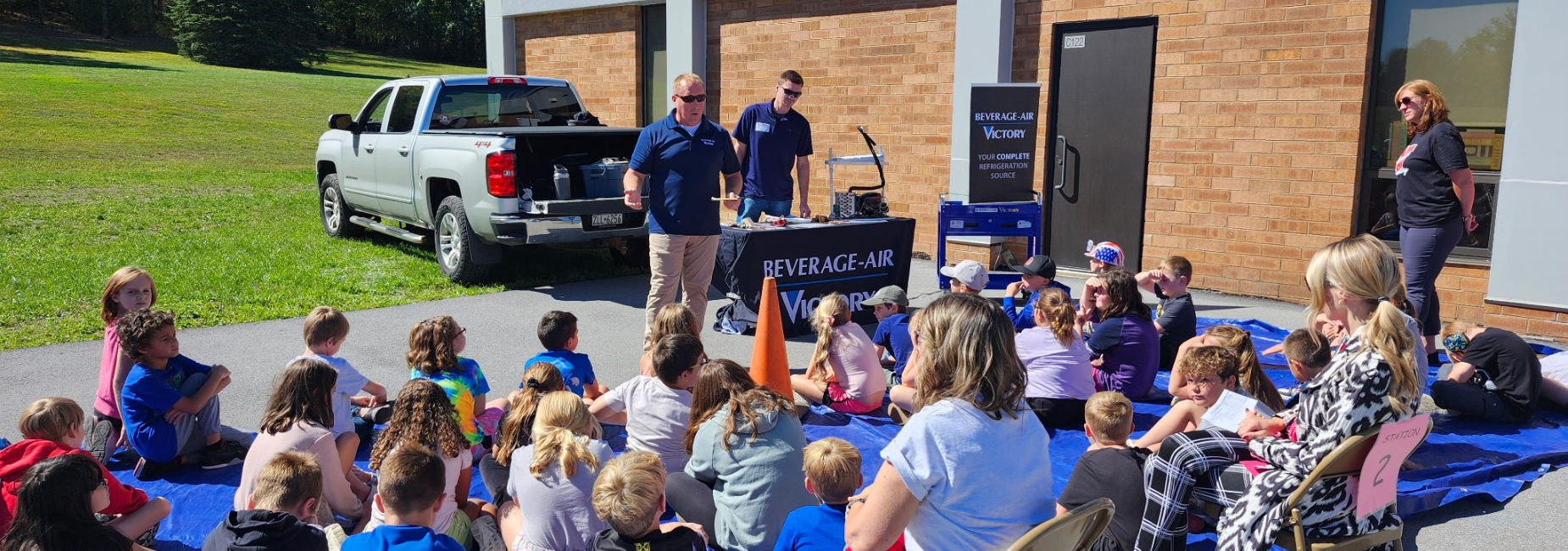 Group of children gathered around two presenters by a pickup truck near a building.