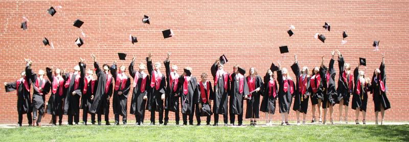 Group of graduates in caps and gowns throwing their hats in celebration against a brick wall.