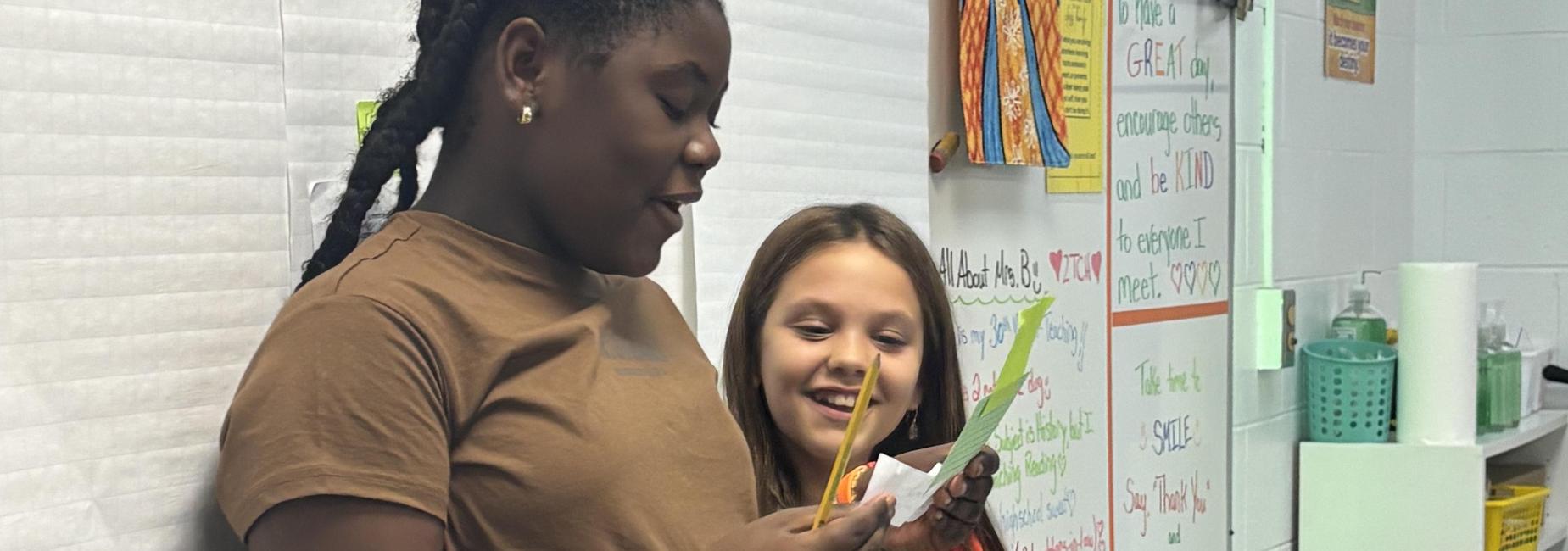 Two girls looking at a paper with a pencil in hand, surrounded by classroom materials.