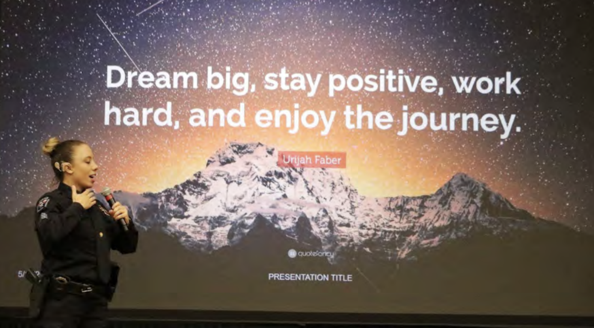 Dalhart Police Officer Erica Trevino stands on stage holding a microphone, delivering a presentation. Behind her, a large screen displays a motivational quote by Urijah Faber that reads, 'Dream big, stay positive, work hard, and enjoy the journey,' set against a mountainous background under a starry sky. Officer Trevino appears to be engaging with the audience, creating an inspiring atmosphere in line with the theme of the presentation.