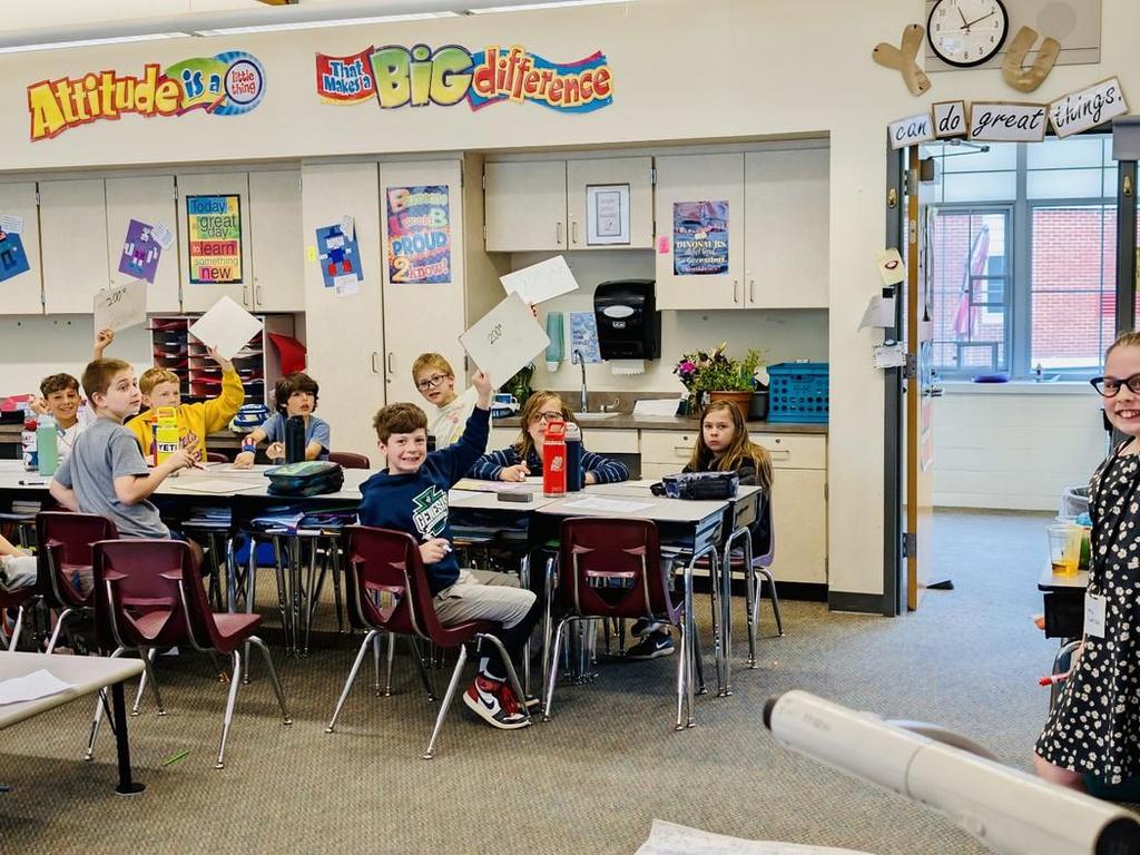 Elementary students in a classroom raising their hands and holding up papers while an elementary student stands at the whiteboard playing teacher.