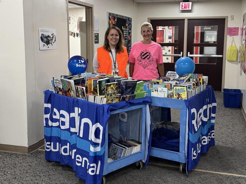 Two women in bright shirts standing behind a book cart filled with books in a hallway.