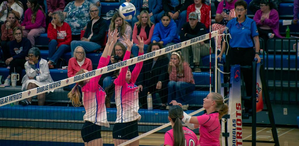 Douglas girls Volleyball players jumping to block a serve in an indoor game setting.