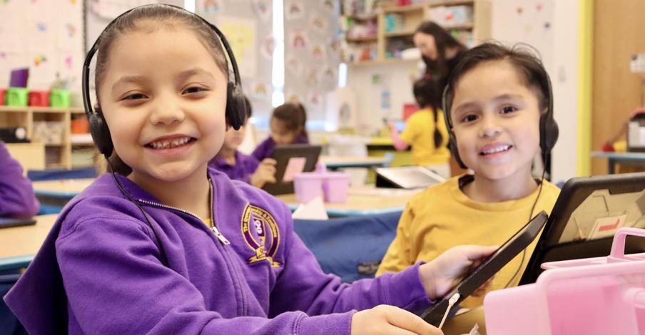 Two children smiling while wearing headphones and using tablets in a classroom setting.