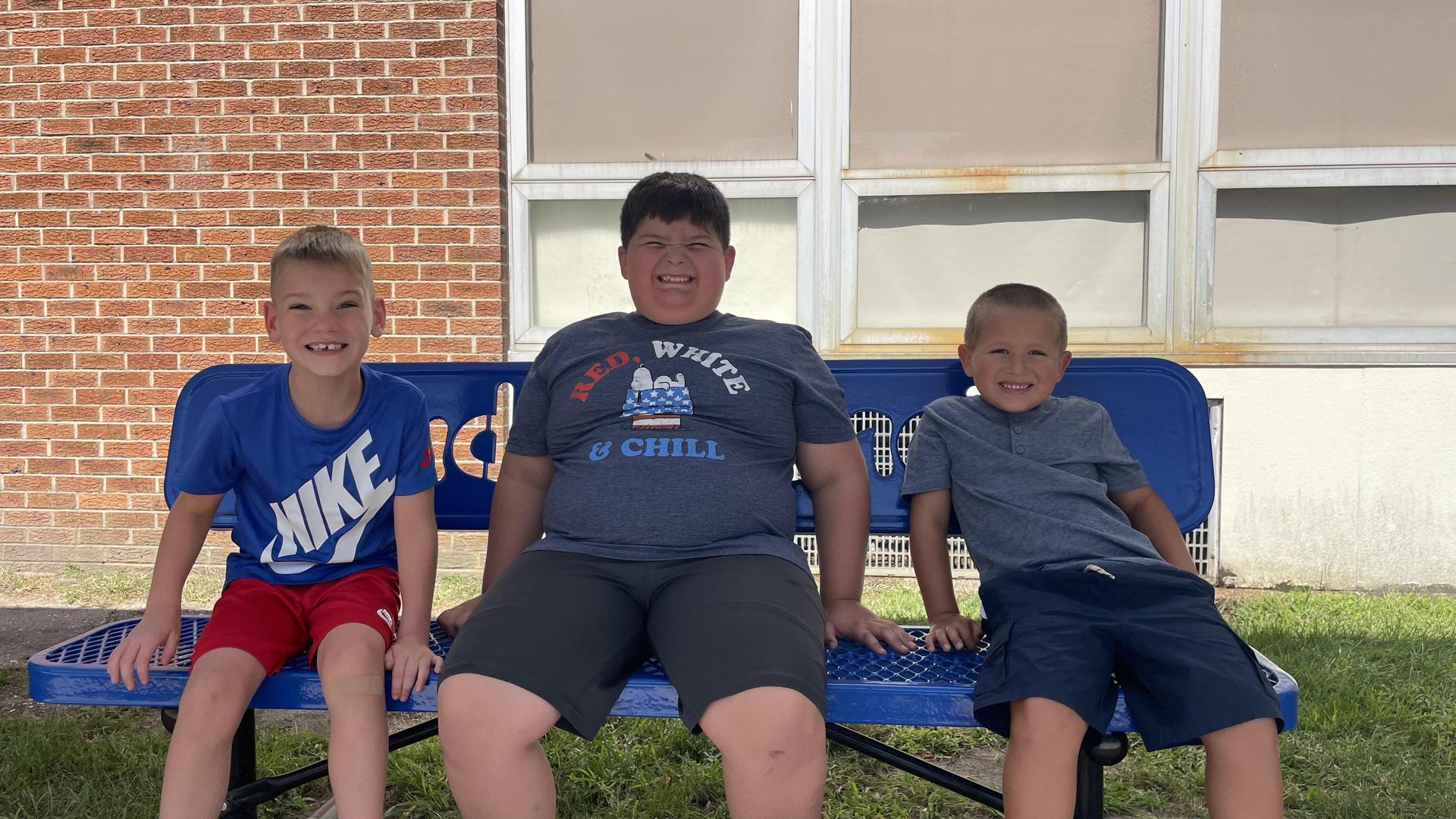 Three smiling boys sitting on a blue bench outside a school building.