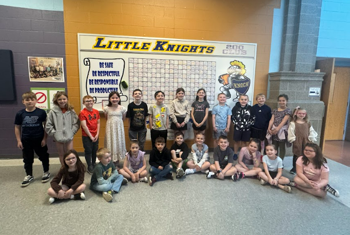 primary students seated and standing smiling at camera in front of knight board