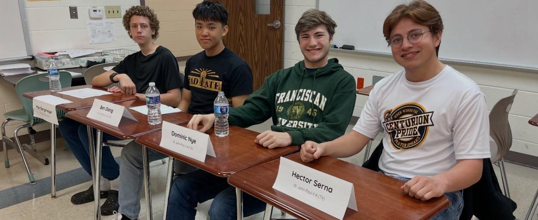 Four students seated at desks with water bottles and name signs in a classroom.