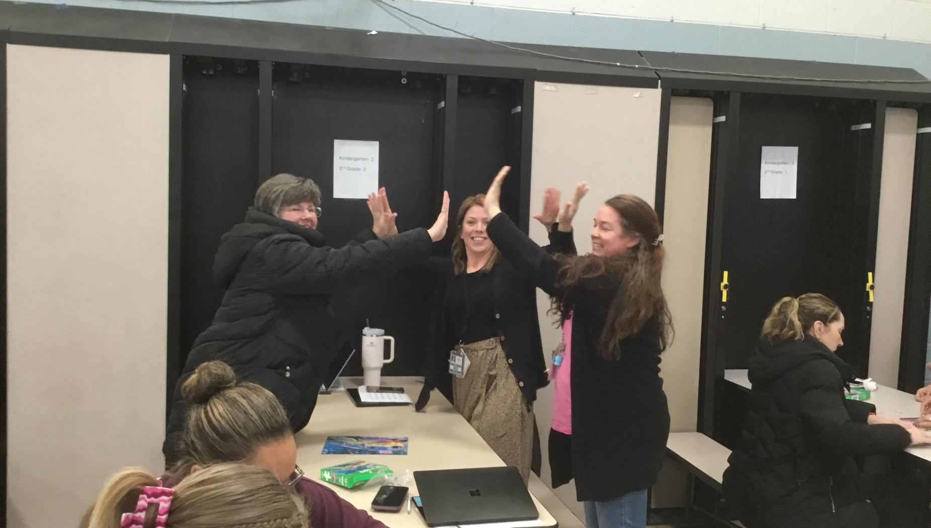 Four women high-fiving in a cafeteria setting, smiling and enjoying the moment.