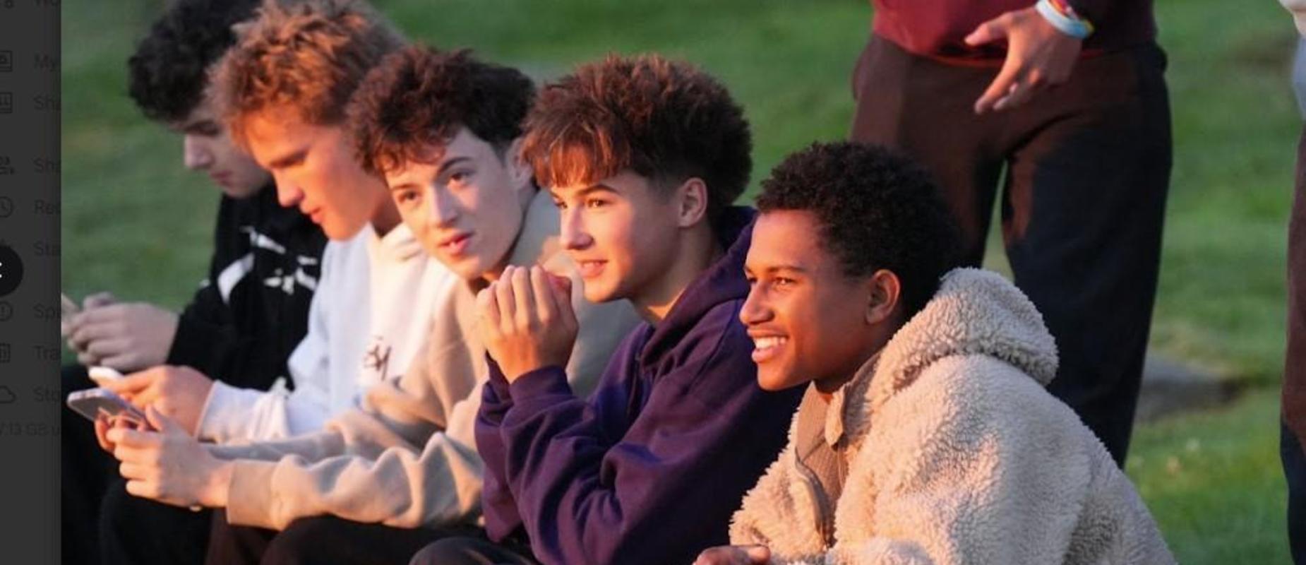 A group of five young men sitting closely together outdoors, smiling and engaged in conversation.