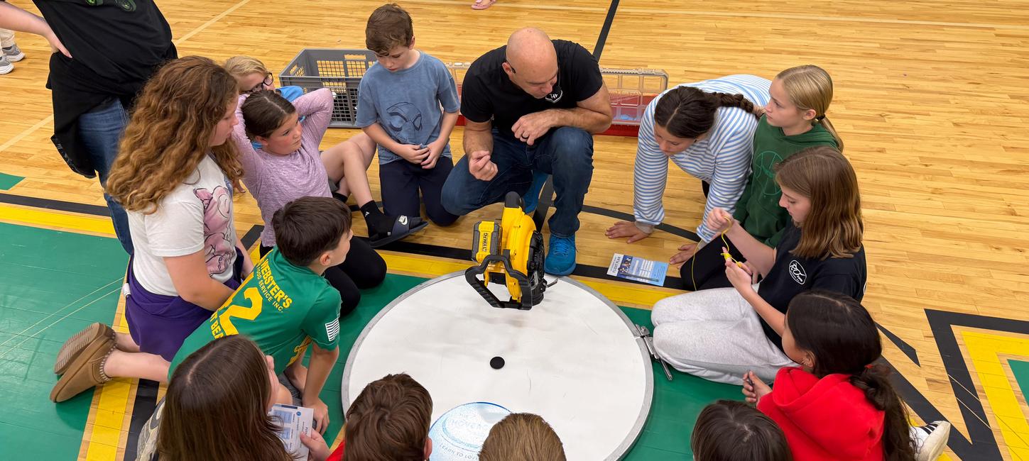 A group of students lean in to hear a teachers instructions on the gym floor