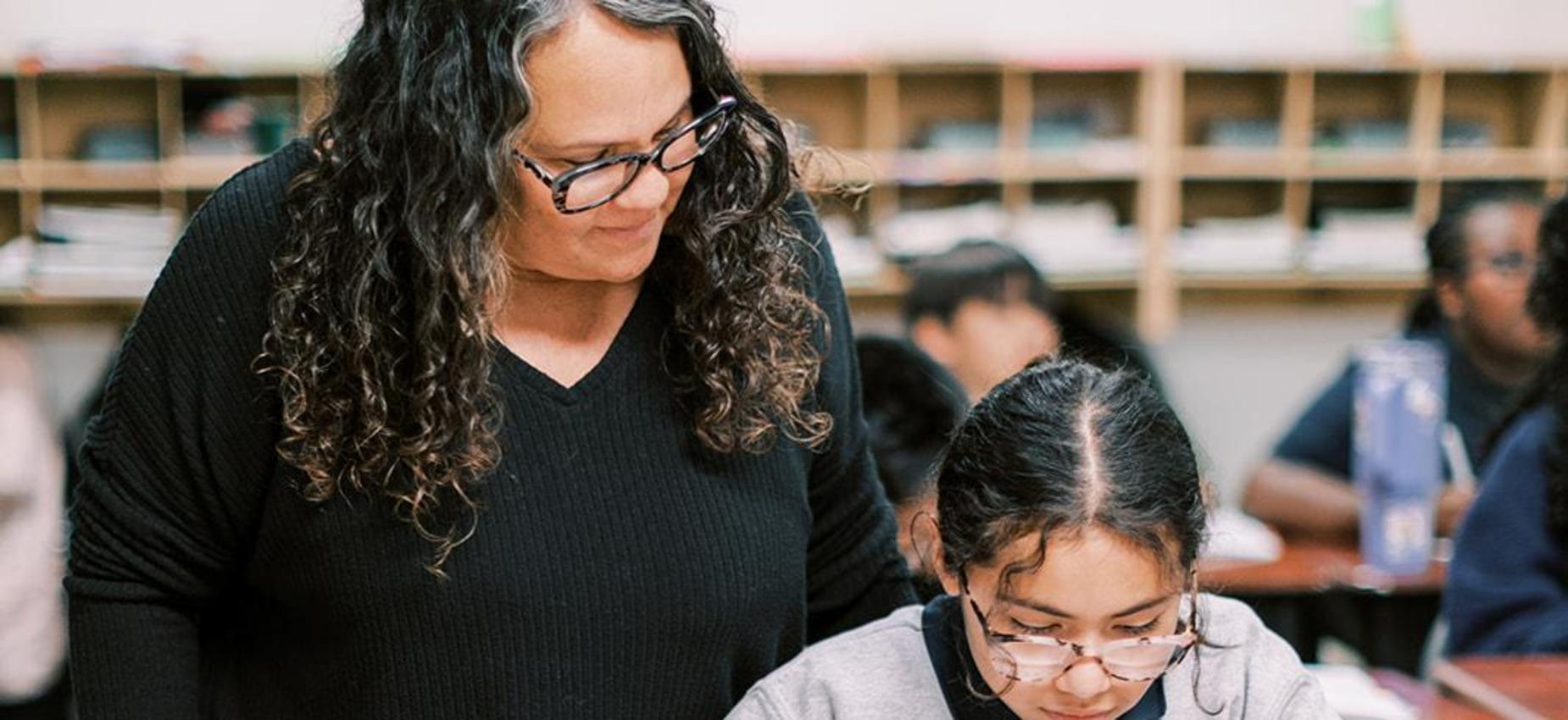 Teacher looks on as a student in a Founders Classical Academy sweatshirt writes at her desk