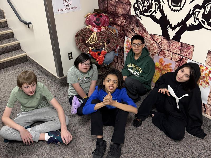 Group of five children sitting on the floor near a painted wall and a scarecrow decoration.