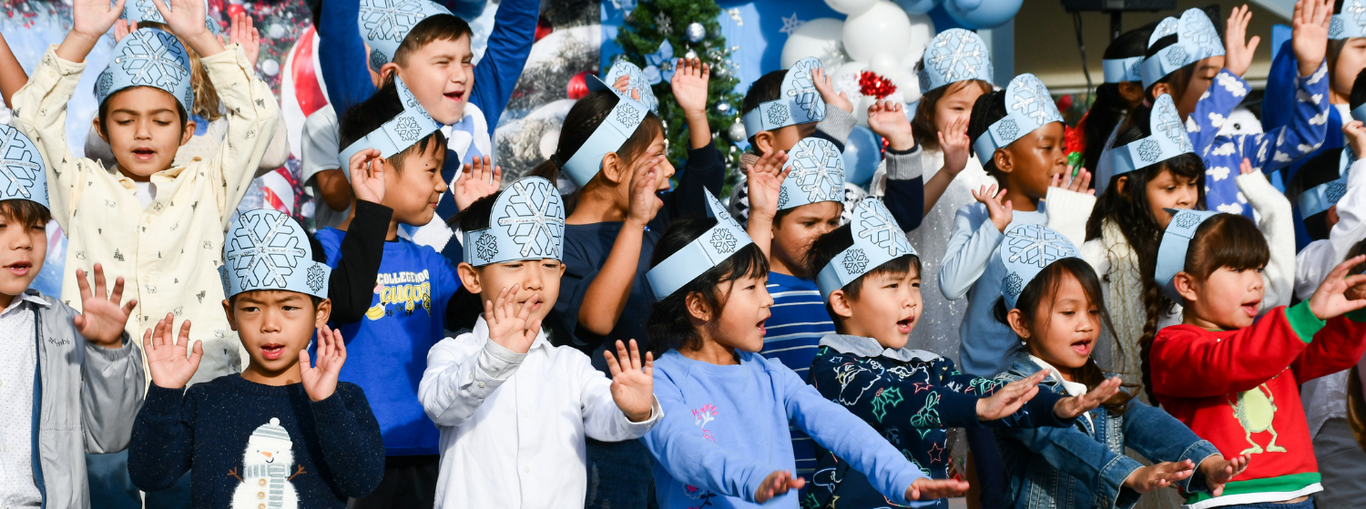 Children in festive hats singing joyfully with festive decorations behind them.
