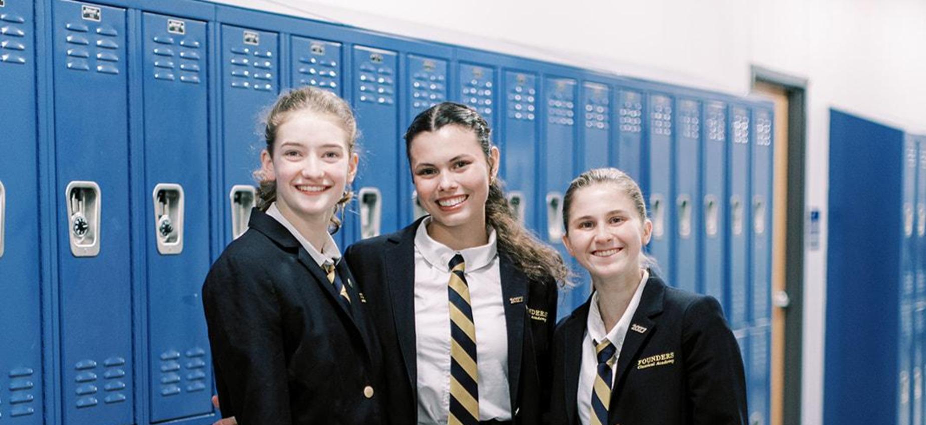 Three smiling high school students in blazers and plaid skirts stand by the lockers