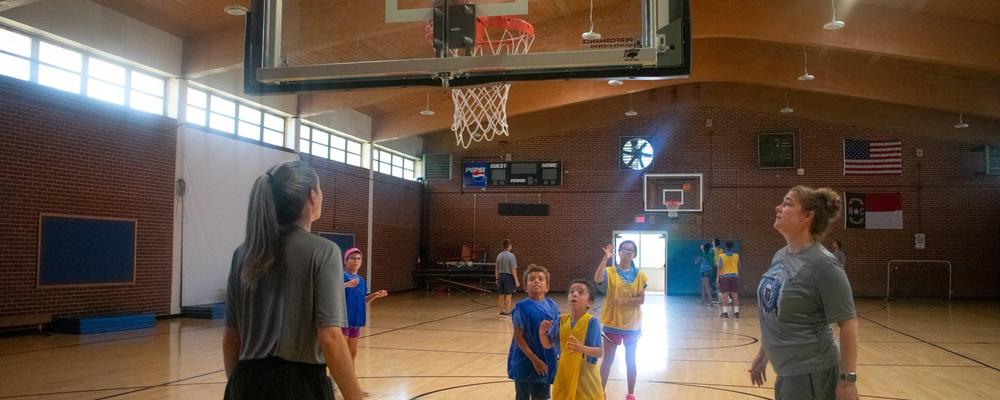 Children playing basketball in a gym with a hoop in the background.