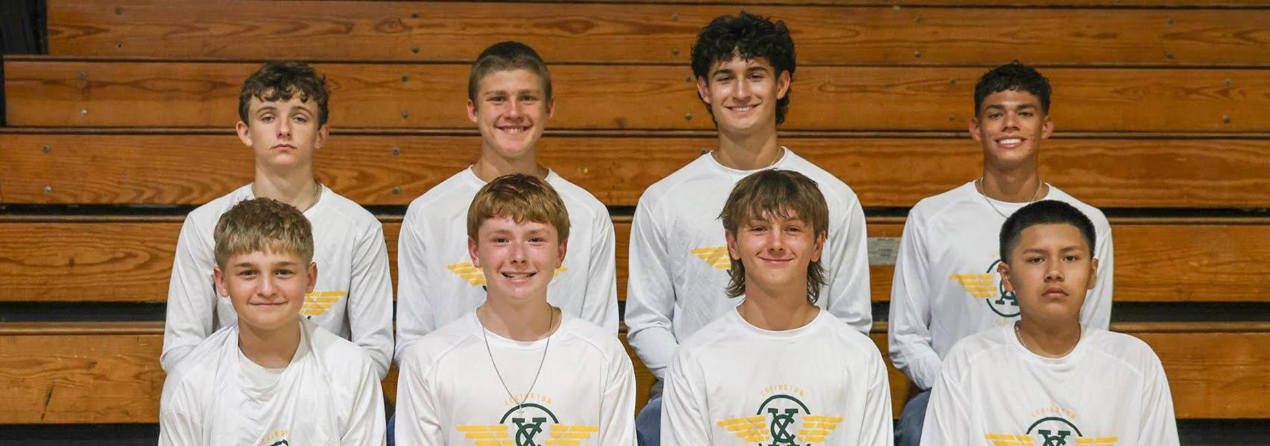 Group of young male athletes in white shirts sitting on gym bleachers.