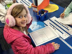 A smiling first grader wearing ear muffs sits at a table in front of a handwritten letter