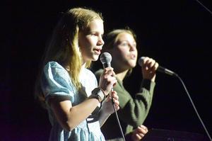 Two girls singing into microphones, one in a blue dress and the other in a sweater.