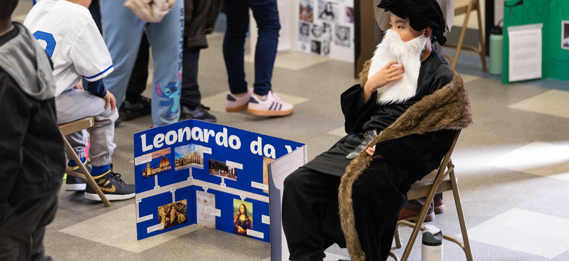 Child dressed as Leonardo da Vinci sitting next to a display board.