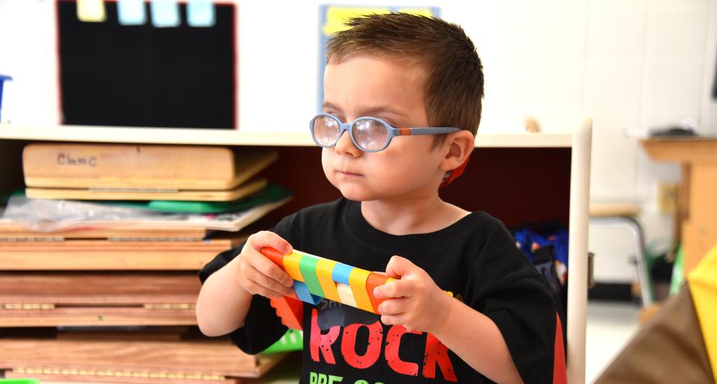 A young child wearing glasses holds a colorful toy in a classroom setting.