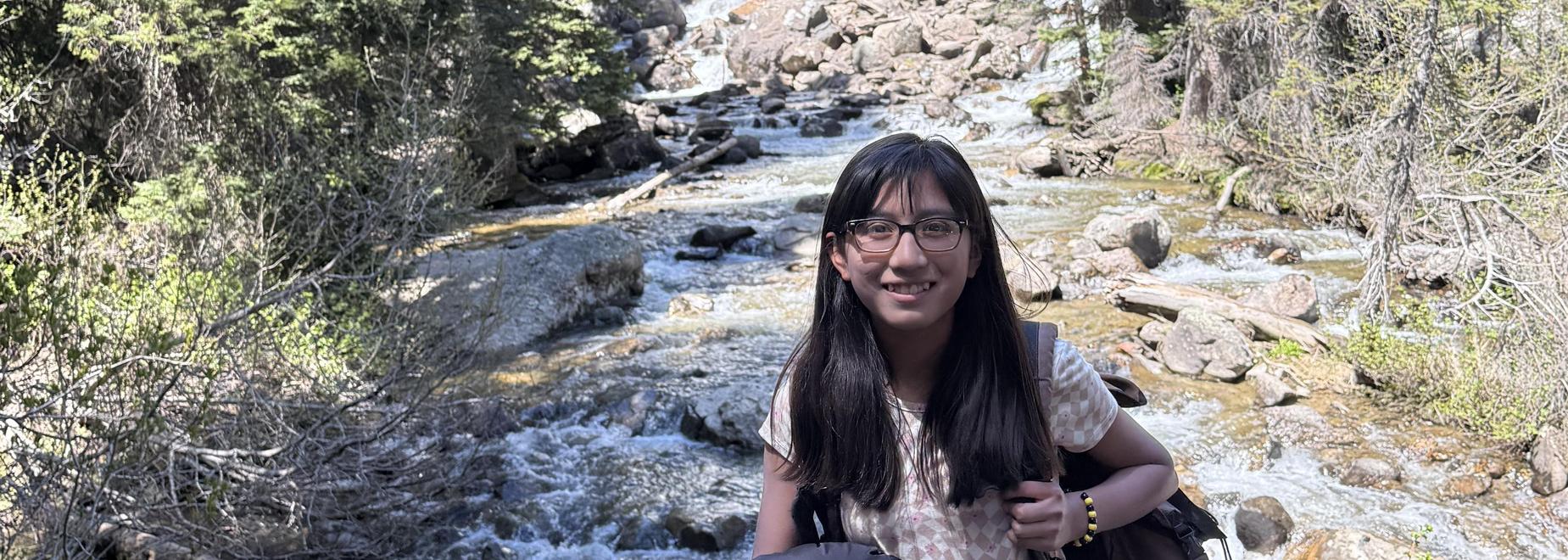 A young woman stands in front of a waterfall surrounded by trees.