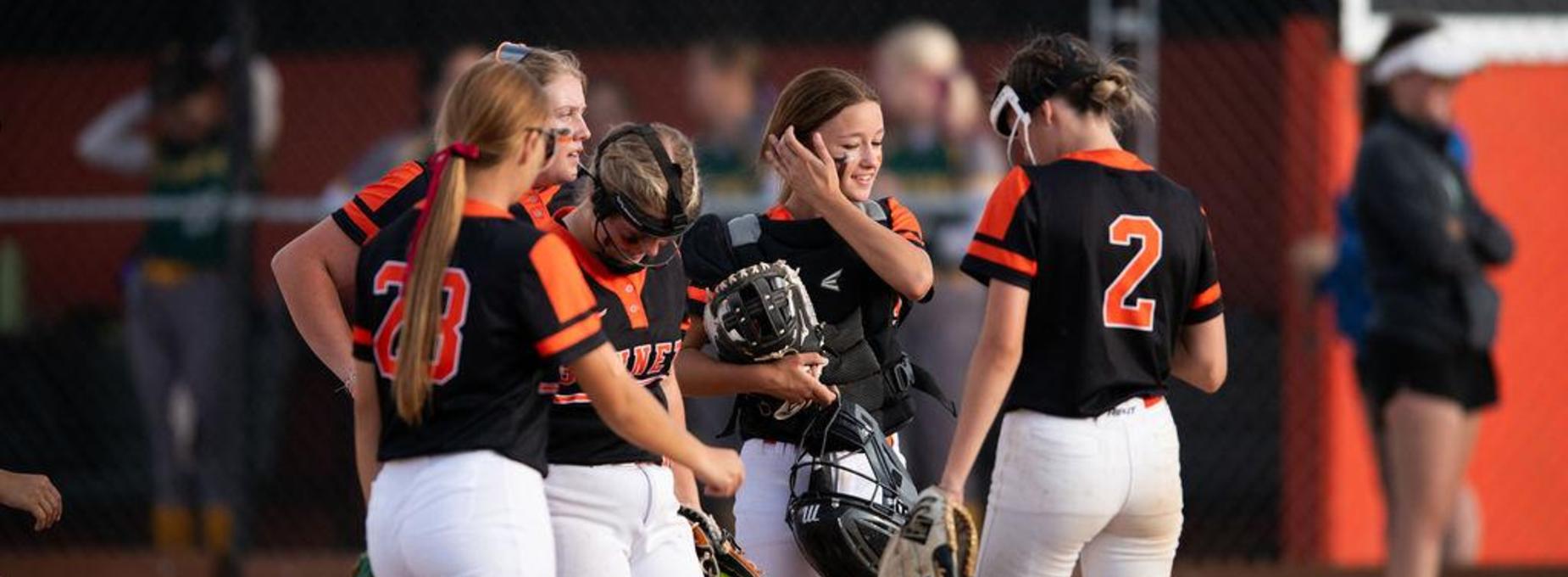 A group of five young female softball players huddle together on the field, discussing the game.
