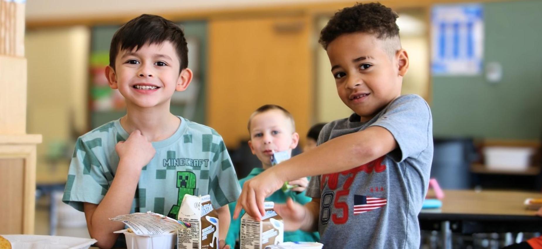 Two boys smiling with drinks, another boy peeking behind them in a casual setting.