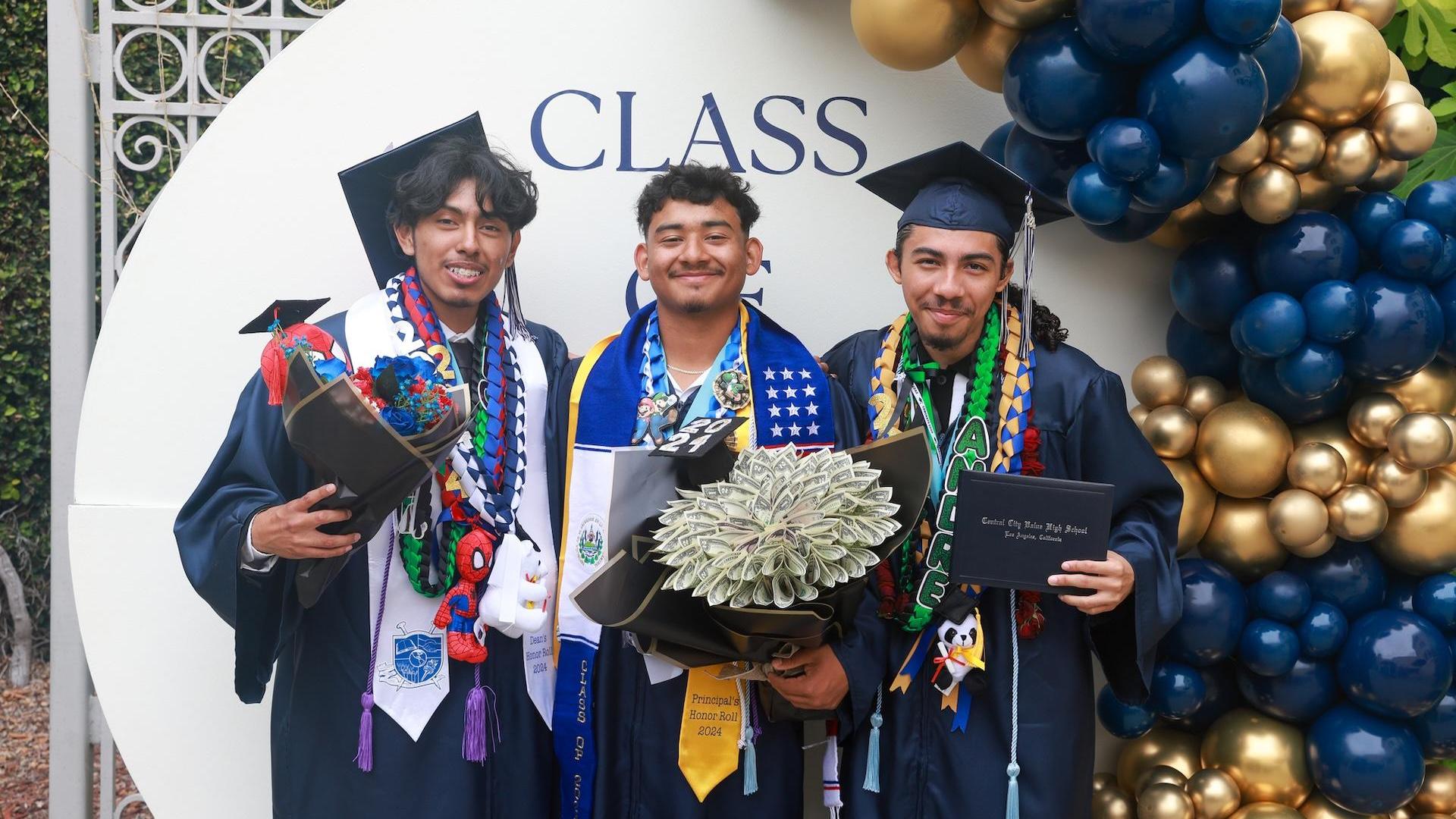 Three graduates in caps and gowns posing with diplomas and flowers.