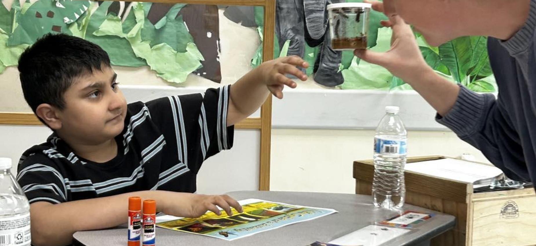 Child reaching for a jar held by an adult in a classroom setting.