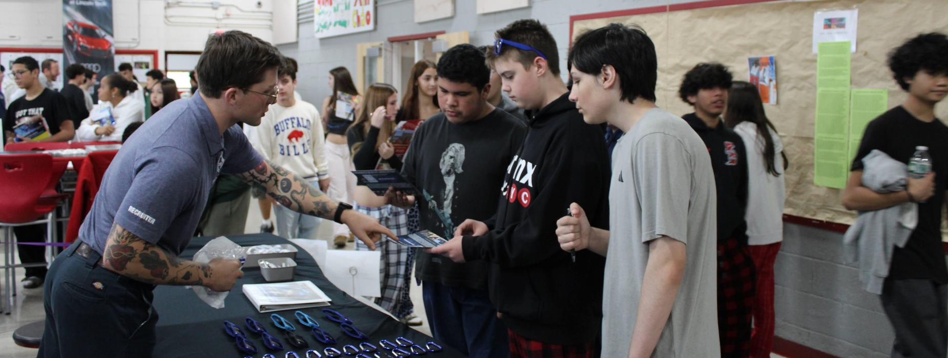 Small group of high school students at the CHSW Career and College Expo lean in and listen to a college recruiter at a table speaking about their school.