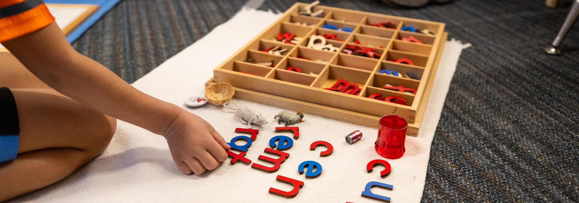 Child's hand arranging colorful letters on a mat beside a wooden letter box.