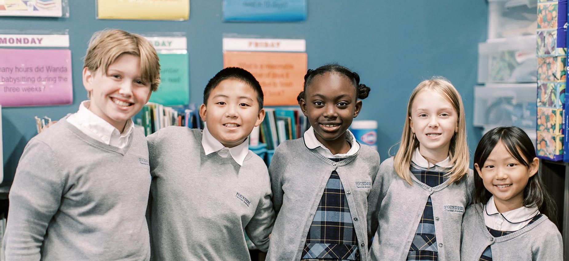 Five smiling students in gray sweaters stand together in a classroom