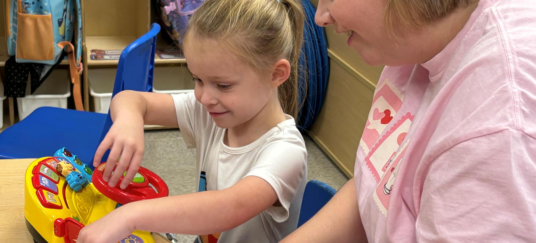 A young girl plays with a colorful toy as a woman assists her, both smiling.