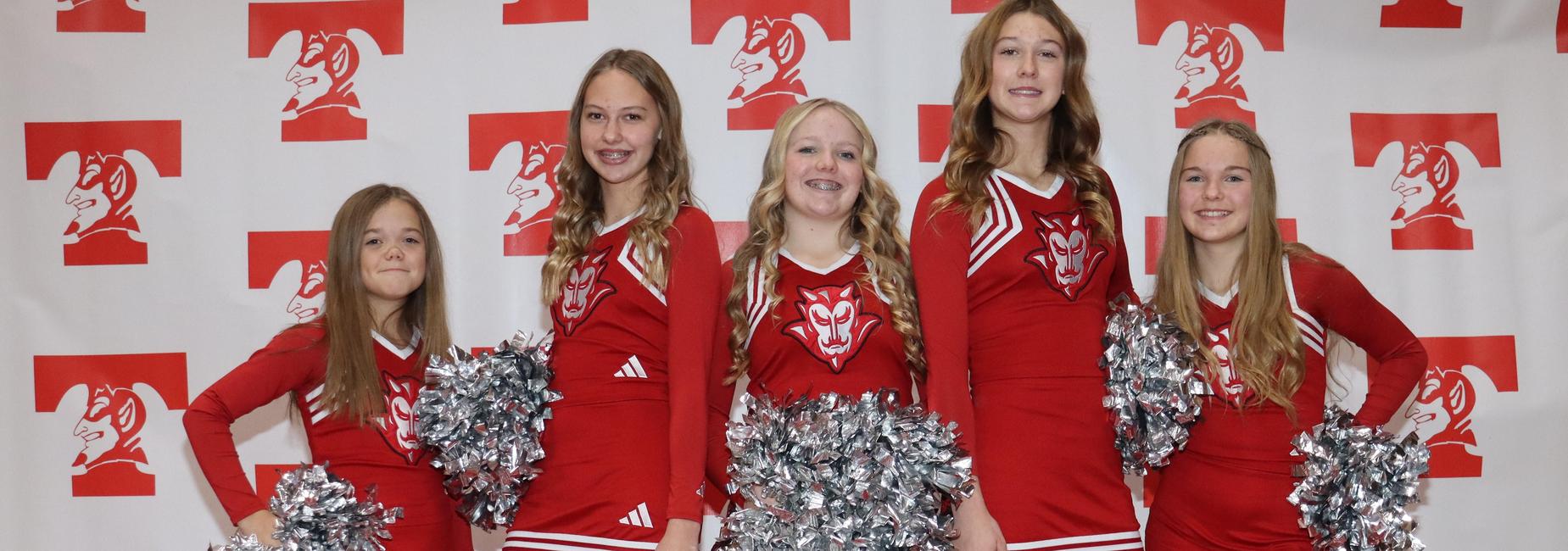 Five cheerleaders in red uniforms with pom-poms posing together on a backdrop.