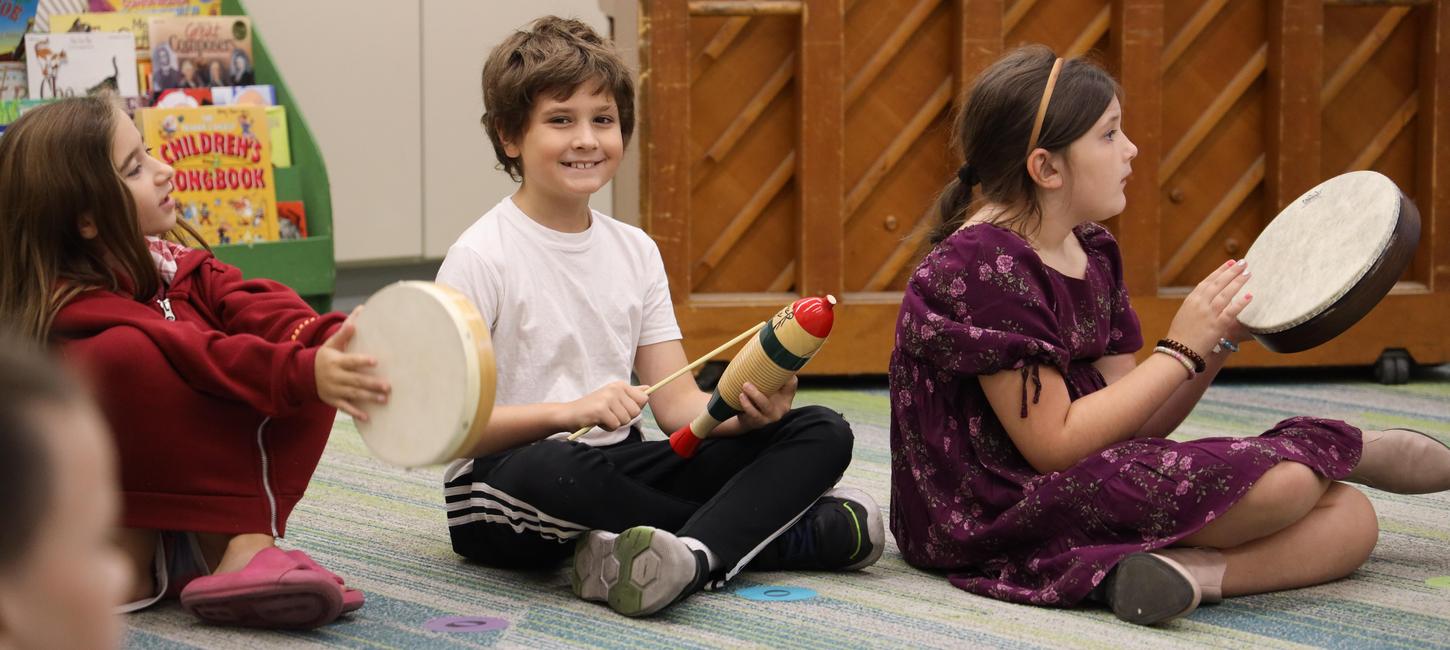 Students smile while sitting on the floor and playing instruments